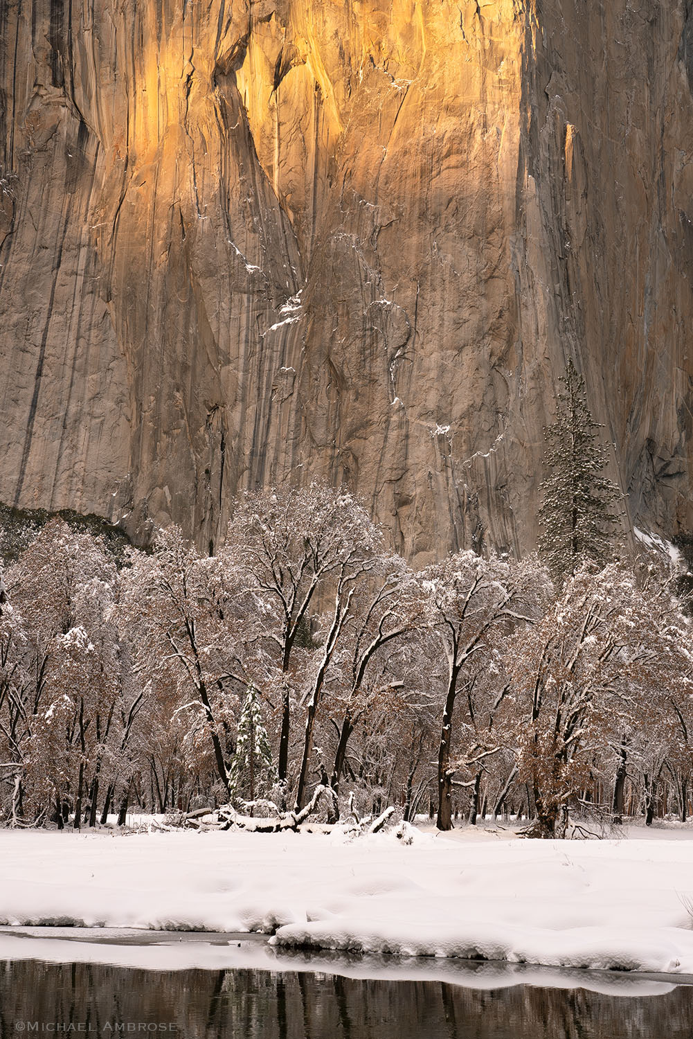The Merced River stands still in front of the "heart" of Iconic El Capitan , which is lit with warm light on a snowy, Yosemite National Park evening.